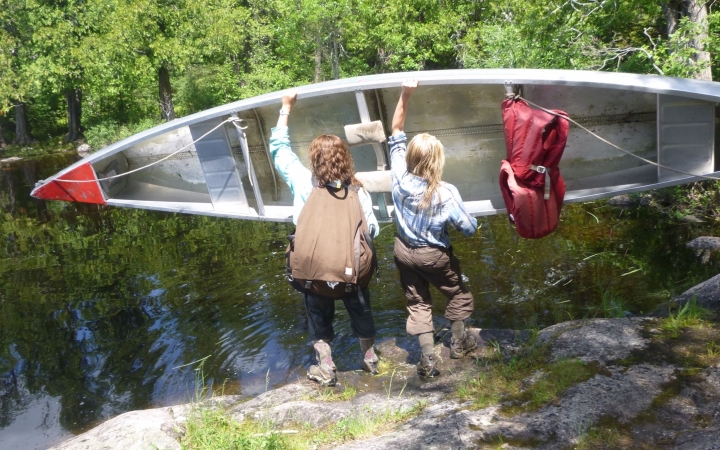 Two people hold a canoe in midair, preparing to lower it into the body of water below them. 